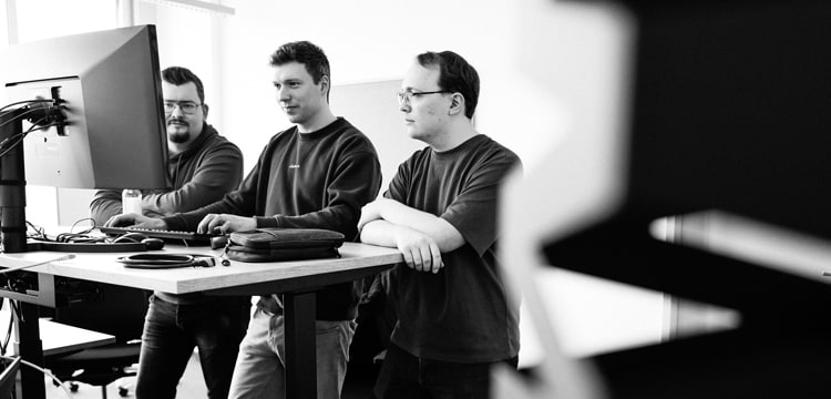 Drei junge Männer stehen an einem Schreibtisch und blicken auf einen Monitor