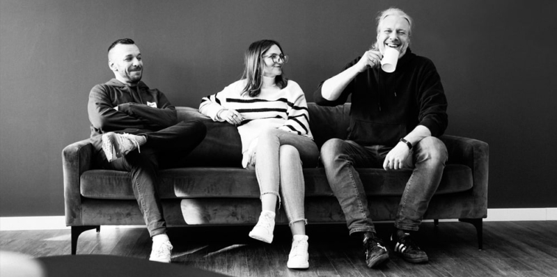 Auf einem Sofa sitzen drei Personen, zwei Personen (Mann und Frau) sehen einen Mann an, der einen Kaffeebecher zum Mund führt und lacht.