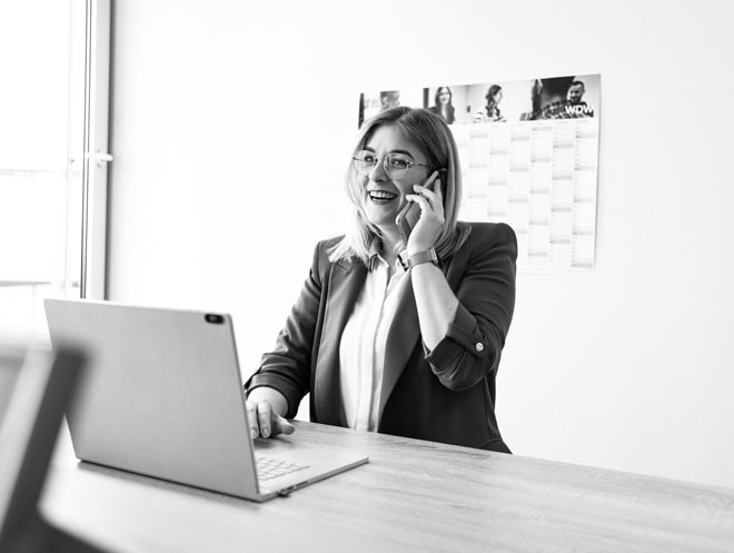 Eine Frau steht vor einem Schreibtisch mit Laptop, hinter ihr hängt ein Kalender. Sie lächelt freundlich während sie mit dem Smartphone telefoniert.