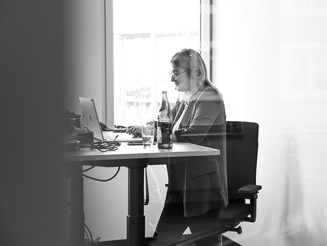 Eine Frau mit Headset auf dem Kopf steht an einem hochgefahrenen Schreibtisch und blick auf ein aufgeklapptes Laptop. Neben diesem steht eine Sprudelflasche und ein halbvolles Glas.