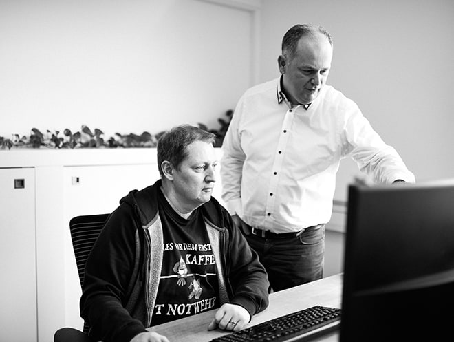 Zwei Männer sehen konzentriert auf einen Bildschirm. Einer der beiden sitzt vor der Tastatur, er trägt eine Sweatshirt Jacke und ein T-shirt mit großem Aufdruck, der andere Mann steht neben ihm, trägt ein weißes Hemd und zeigt auf den Bildschirm.