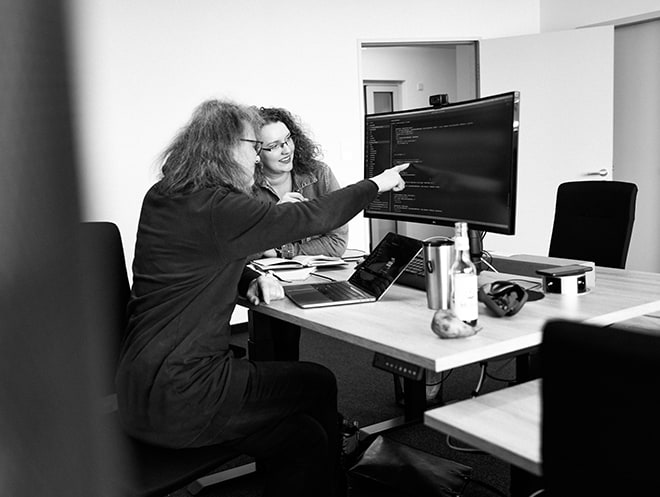 Ein Mann und eine Frau sitzen an einem Schreibtisch. Vor ihnen ist ein großer Bildschirm mit Codezeilen zu sehen, sowie ein aufgeklapptes Laptop. Der Mann zeigt mit dem Finger auf den Bildschirm.