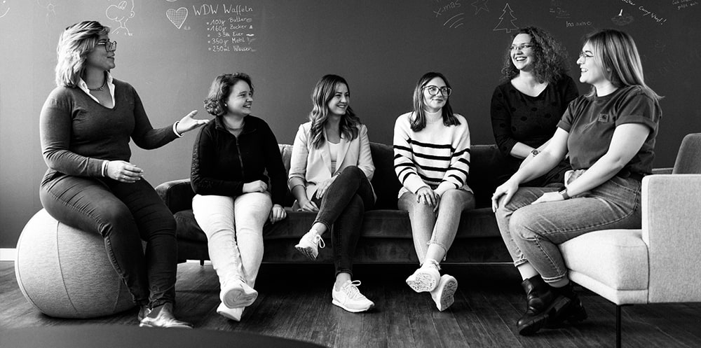 Auf dem Bild sind 6 Frauen zu sehen, die sich anlächeln. 4 sitzen auf einem Sofa, eine auf einem Sitzball und eine auf einem Sessel. Sie scheinen im Gespräch zu sein und lachen über etwas. Hinter ihnen ist eine Tafelwand mit Zeichnungen und einem Waffelrezept zu sehen.