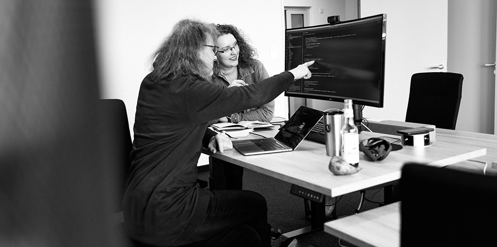 Ein Mann und eine Frau sitzen an einem Schreibtisch. Vor ihnen ist ein großer Bildschirm mit Codezeilen zu sehen, sowie ein aufgeklapptes Laptop. Der Mann zeigt mit dem Finger auf den Bildschirm.