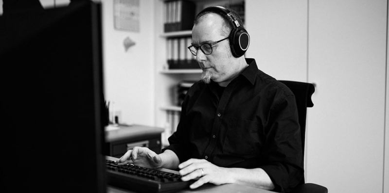 Ein mann mit großen Kopfhörern auf den Ohren sitzt in einem Büro vor einer Tastatur und blickt konzentriert auf diese.