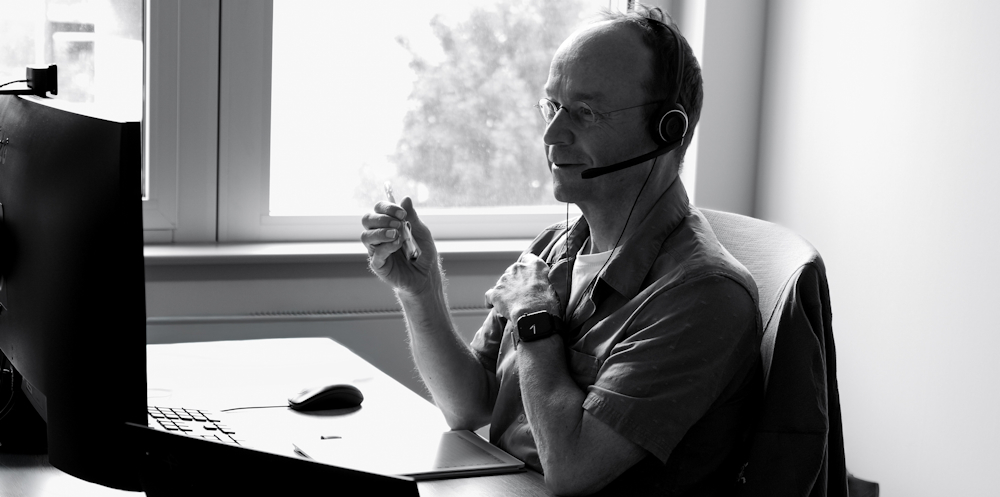 Ein Mann sitzt an einem Schreibtisch vor einem Monitor und führt mit Headset ein Gespräch. Er hält einen Stift in der Hand und wirkt konzentriert. Das Schwarz-Weiß-Foto zeigt ihn seitlich vor einem Fenster sitzend.