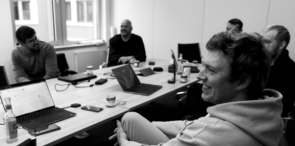 Mehrere Personen sitzen entspannt in einem Besprechungsraum an einem Tisch mit Laptops und Getränken. Sie unterhalten sich und lachen, die Szene wirkt locker und kollegial. Das Schwarz-Weiß-Foto zeigt die Gruppe aus einer seitlichen Perspektive.