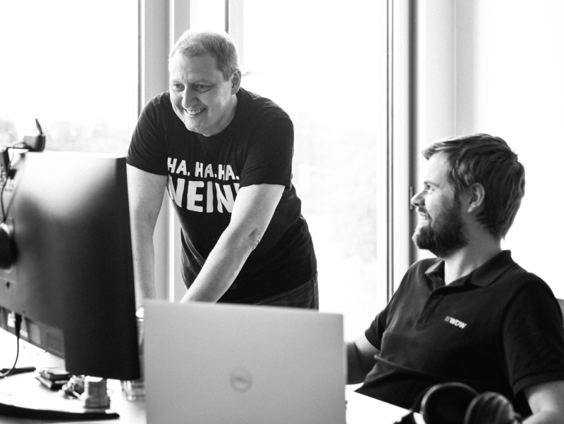 Zwei Männer stehen bzw. sitzen gemeinsam an einem Schreibtisch vor Monitoren und lachen. Der stehende Mann trägt ein T-Shirt mit dem Aufdruck ‚HA. HA. HA. NEIN.‘, der sitzende Mann schaut ihn lächelnd an. Das Schwarz-Weiß-Foto zeigt eine freundliche, lockere Arbeitssituation.
