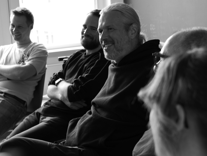 Mehrere Personen sitzen nebeneinander in einem Besprechungsraum und lachen. Die Gruppe wirkt entspannt und aufmerksam, während sie einem Gespräch oder einer Präsentation folgt. Das Schwarz-Weiß-Foto zeigt die Personen seitlich im Profil.