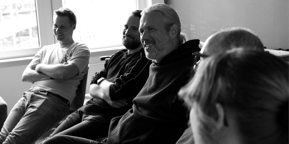 Mehrere Personen sitzen nebeneinander in einem Besprechungsraum und lachen. Die Gruppe wirkt entspannt und aufmerksam, während sie einem Gespräch oder einer Präsentation folgt. Das Schwarz-Weiß-Foto zeigt die Personen seitlich im Profil.
