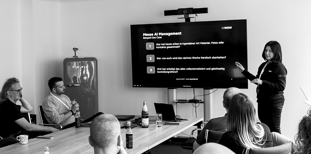 Eine Frau steht in einem Besprechungsraum vor einem Bildschirm und präsentiert einen Anwendungsfall zum Thema AI Management. Mehrere Personen sitzen um einen Tisch und verfolgen die Präsentation. Die Szene ist in Schwarz-Weiß aufgenommen.